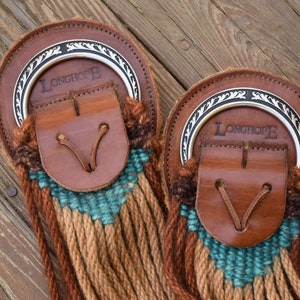 May include: Two leather and woven horse tack pieces. Each piece has a brown leather backing with a silver metal ring and the word "LONGHOPE" printed on it. The woven section features teal, tan, and brown colors.