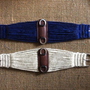 K&ouml;nnte beinhalten: Zwei Pferdegurte. Einer ist marineblau, der andere cremefarben. Beide haben ein gewebtes Design, eine Leder-Mitte und silberfarbene Metallbeschl&auml;ge. Die Gurte dienen dazu, einen Sattel auf einem Pferd zu befestigen.