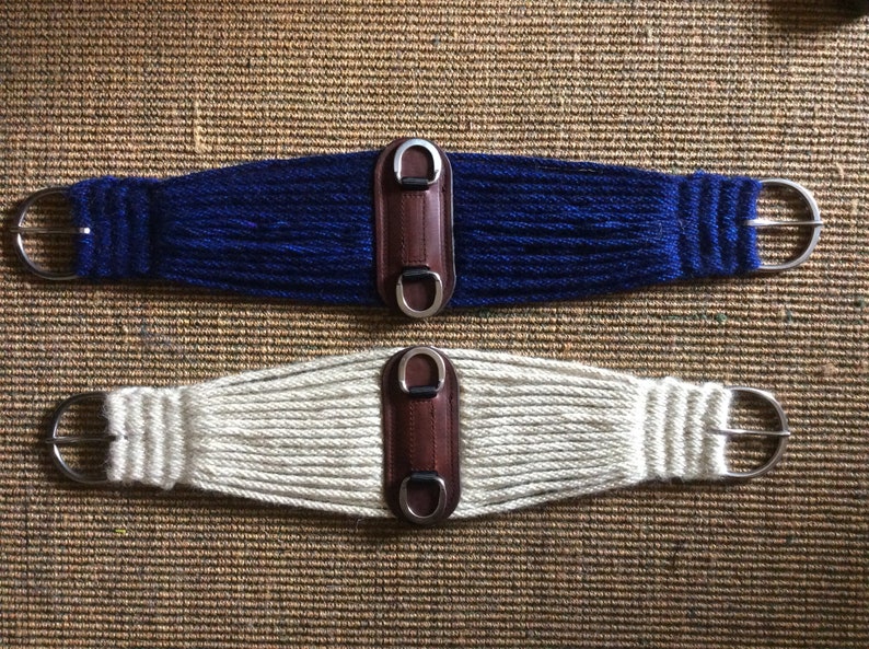 K&ouml;nnte beinhalten: Zwei Pferdegurte. Einer ist marineblau, der andere cremefarben. Beide haben ein gewebtes Design, eine Leder-Mitte und silberfarbene Metallbeschl&auml;ge. Die Gurte dienen dazu, einen Sattel auf einem Pferd zu befestigen.