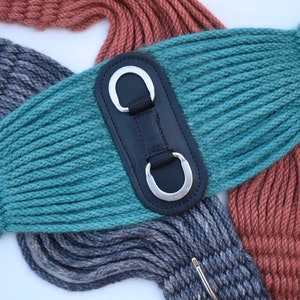 K&ouml;nnte beinhalten: Nahaufnahme von drei Pferdegurten in den Farben Teal, Grau und Rost. Der teal-farbene Gurt hat ein schwarzes Leder-Mittelst&uuml;ck mit zwei silbernen D-Ringen. Die grauen und rostfarbenen Gurte sind im Hintergrund zu sehen.