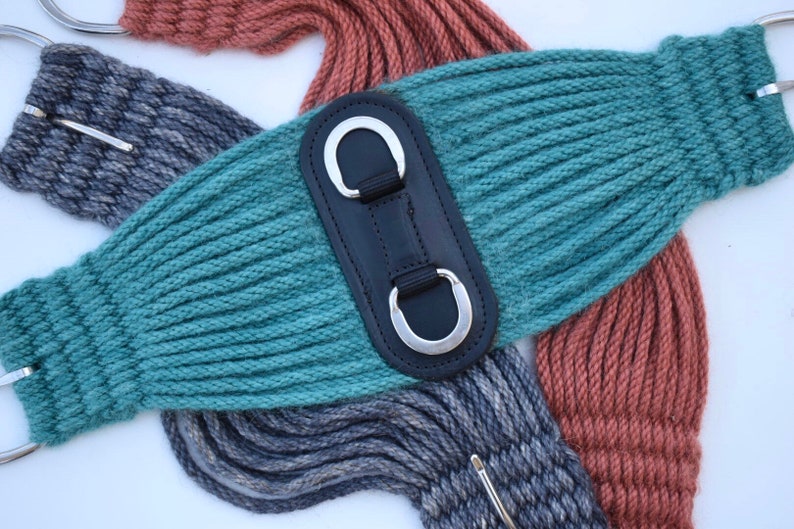 K&ouml;nnte beinhalten: Nahaufnahme von drei Pferdegurten in den Farben Teal, Grau und Rost. Der teal-farbene Gurt hat ein schwarzes Leder-Mittelst&uuml;ck mit zwei silbernen D-Ringen. Die grauen und rostfarbenen Gurte sind im Hintergrund zu sehen.