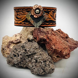 May include: A wide leather cuff bracelet featuring a tan and black tooled floral pattern. The center is adorned with a diamond-shaped metal plate and a layered copper flower embellishment with a textured silver-toned center stud detail.
