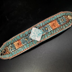 May include: A turquoise leather bracelet with intricate embossed patterns and decorative elements. The bracelet features squares of orange and blue, with a square white charm in the center. Gold-colored studs and a brass button complete the design.
