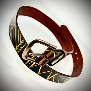 May include: A brown leather belt featuring a dark green painted section with gold geometric line patterns, including chevrons and cross-hatching. The belt is equipped with a rectangular, weathered brass-toned metal buckle and a single leather keeper.