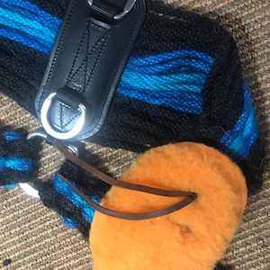 K&ouml;nnte beinhalten: Nahaufnahme einer schwarzen und blauen gewebten Pferdesattelunterlage mit einem schwarzen Lederriemen und silbernen Metallringen. Ein orangefarbenes, flauschiges Pad mit braunen Lederriemen ist befestigt.