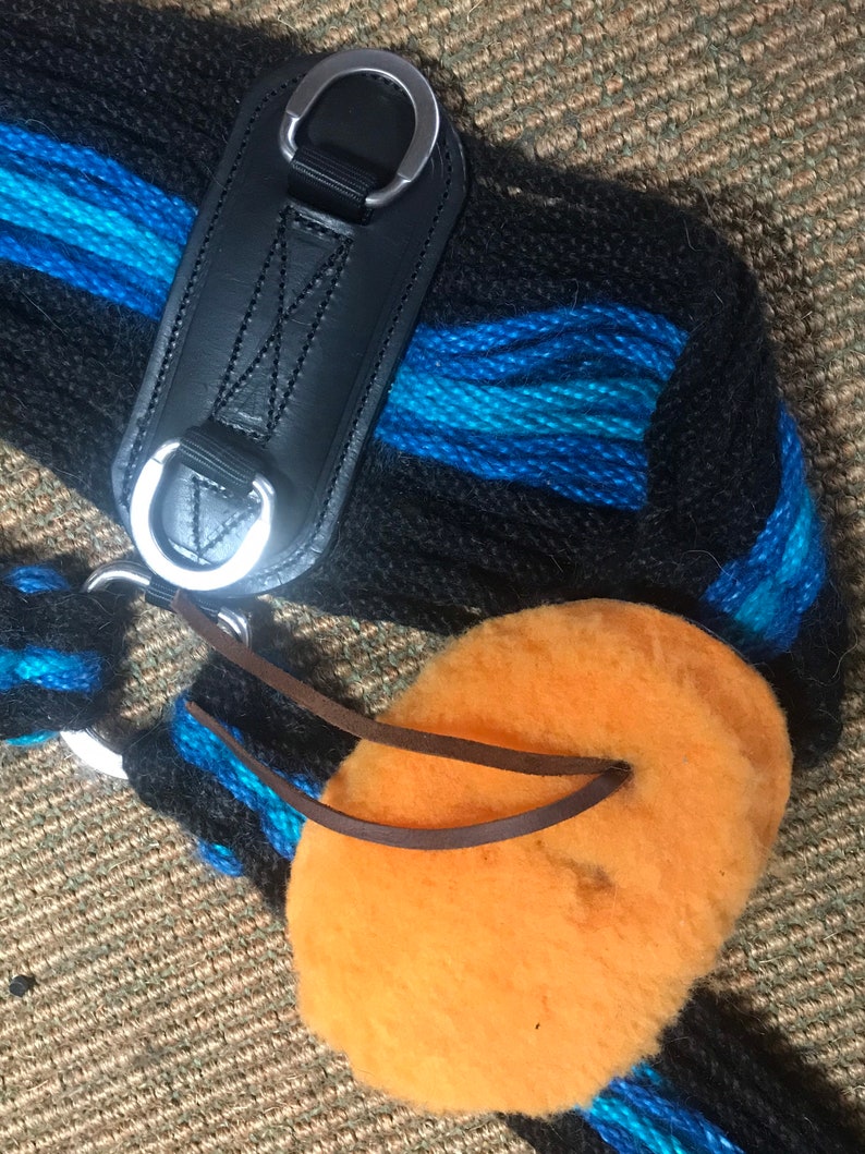 K&ouml;nnte beinhalten: Nahaufnahme einer schwarzen und blauen gewebten Pferdesattelunterlage mit einem schwarzen Lederriemen und silbernen Metallringen. Ein orangefarbenes, flauschiges Pad mit braunen Lederriemen ist befestigt.