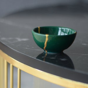 May include: A small, green bowl with gold accents sits on a black marble table with a gold metal base.