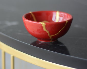 May include: A small red bowl with gold cracks. The bowl is sitting on a black marble surface.
