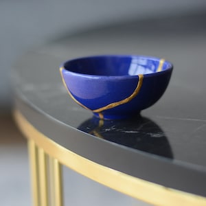 Könnte beinhalten: Eine kleine blaue Keramikschüssel mit goldener Craquelé-Glasur. Die Schüssel steht auf einem schwarzen Marmortisch mit goldenem Rand.