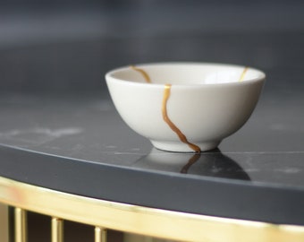 Peut inclure: Un petit bol en céramique blanc cassé avec une ligne de réparation dorée. Le bol est posé sur une surface gris foncé avec une bordure en métal doré. L'arrière-plan est flou.