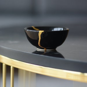 May include: A small black ceramic bowl with a gold crackle design. The bowl is sitting on a black and gold table.
