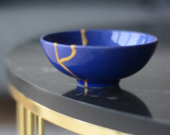 May include: A small blue ceramic bowl with gold crackle lines. The bowl is sitting on a black marble table with a gold metal base.