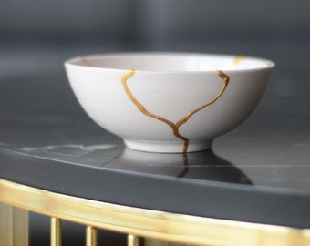 Peut inclure: Un bol en céramique blanche avec des détails craquelés dorés posé sur une table en marbre noir avec une base en métal doré.