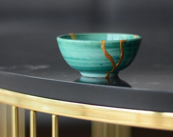 May include: A small, turquoise ceramic bowl with gold kintsugi repair lines. The bowl sits on a dark surface, with a gold metal frame in the foreground. The bowl's design reflects the Japanese art of embracing imperfections, a beautiful piece.