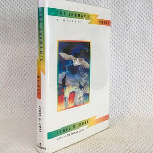 May include: A hardcover book titled "THE SHAMAN'S BONES" by James D. Doss. The cover features a colorful illustration of a shaman, an owl, and a horse on a cliffside with lightning. The spine also displays the title and author's name.
