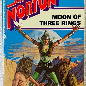 May include: A vintage paperback book cover for "Moon of Three Rings" by Andre Norton. The cover depicts a woman in green and black clothing with her arms raised, surrounded by fantastical creatures against a desert background. The author's name is in large red letters.