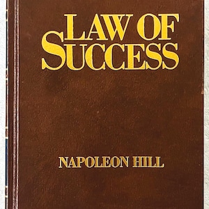 以下が含まれることがあります： ナポレオン・ヒル著『Law of Success』と題された茶色のハードカバー本。表紙には大きく太い黄色のアルファベット大文字でタイトルが記されており、その下部中央には、より小さな黄色のアルファベット大文字で著者名が印刷されています。