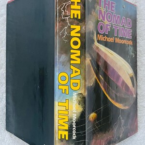 Puede incluir: Un libro de tapa dura titulado "The Nomad of Time" de Michael Moorcock. El lomo es amarillo con el t&iacute;tulo en letras grandes y verticales. La portada presenta una ilustraci&oacute;n de dirigible p&uacute;rpura sobre un fondo oscuro.