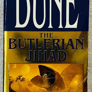 May include: A hardcover book titled "Dune: The Butlerian Jihad" by Brian Herbert and Kevin J. Anderson. The cover features a blue and gold design with the text "The New York Times Bestseller" and an illustration of a desert scene with a large sandworm.