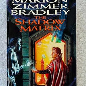 May include: A paperback book cover for "The Shadow Matrix" by Marion Zimmer Bradley. The cover depicts a fantasy scene with two figures, a vibrant sunset, and the book's title in bold orange letters.