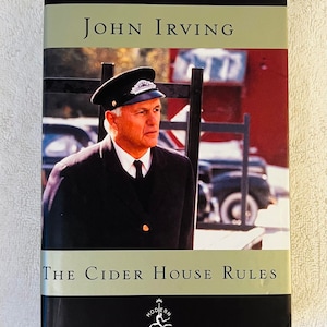 May include: A hardcover book titled "The Cider House Rules" by John Irving. The book cover features a man in a uniform hat and jacket. The Modern Library logo is at the bottom. The book has a black spine and a light green band.