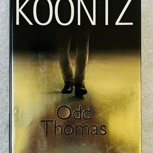 May include: A book cover for "Odd Thomas" by Dean Koontz. The cover features the author's name in white at the top, the title in large letters, and a blurred image of a person's legs and feet. The background is a gradient of gold and black.