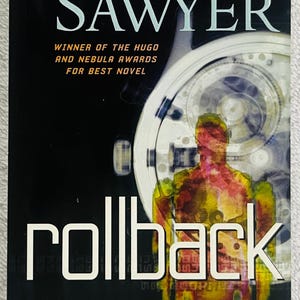 Op de afbeelding: Boekomslag van "Rollback" door Robert J. Sawyer. De titel is wit, met de naam van de auteur erboven. De achtergrond is zwart met een horloge-achtig ontwerp en een figuur in geel en rood.