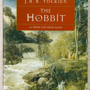 May include: A hardcover book titled "The Hobbit" by J.R.R. Tolkien. The cover features a watercolor painting of a river scene with a stone bridge, trees, and a figure in a long coat. The title is in white letters on a red background.