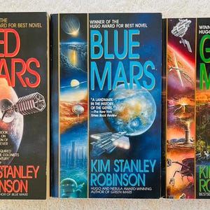 May include: Three science fiction novels by Kim Stanley Robinson are displayed. The covers feature vibrant artwork with titles "Red Mars", "Blue Mars", and "Green Mars". Each book cover includes the author's name and awards.