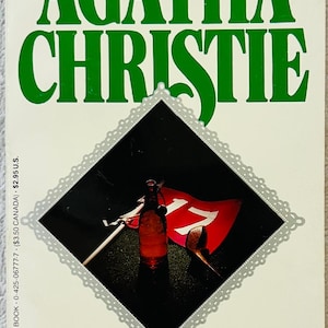 Op de afbeelding: Een paperback getiteld The Boomerang Clue van Agatha Christie. De kaft bevat grote groene tekst en een centrale ruitvormige afbeelding met een golfvlag met het nummer 17, een glazen fles en een pijp. De tekst luidt Mystery's #1 Bestseller.