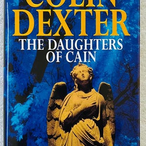 May include: A book cover for "The Daughters of Cain" by Colin Dexter. The title and author's name are in yellow against a blue background. An angel statue is featured on the cover. The book is part of "The New Inspector Morse Novel" series.