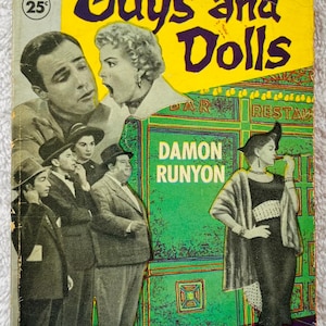 May include: A vintage paperback book cover for "Guys and Dolls." The cover art depicts characters from the story, including a woman in a black dress and a man in a suit. The title is in large purple letters, with the author's name, Damon Runyon, displayed.