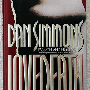 May include: Hardcover book titled "Lovedeath" by Dan Simmons. The cover features a close-up of a person's lips and the title in large, silver lettering. The book is a work of passion and horror.