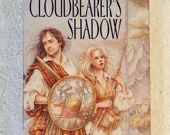 May include: A paperback book titled "Cloudbearer's Shadow" by Ann Marston. The cover art features a man with a shield and a woman with a rifle, set against a backdrop of mountains and a wolf. The book's title is in large, bold letters.