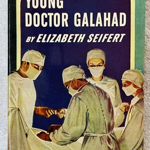 May include: Vintage paperback book cover titled "Young Doctor Galahad" by Elizabeth Seifert. The cover features a dark blue background with white text and an illustration of a surgical scene with four figures in surgical attire. The book is a Pocket Book Edition.