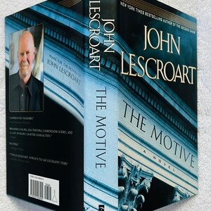 May include: A hardcover book titled "The Motive" by John Lescroart. The cover features a blue and white architectural design with the author's name and book title in gold lettering. A portrait of the author is on the side.