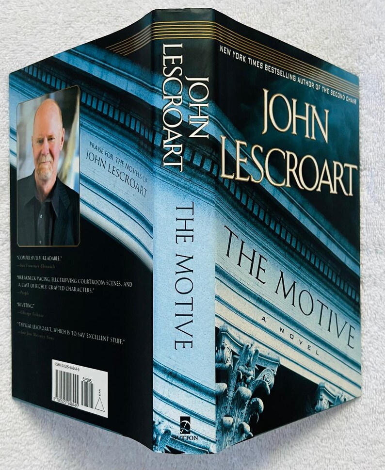 May include: A hardcover book titled "The Motive" by John Lescroart. The cover features a blue and white architectural design with the author's name and book title in gold lettering. A portrait of the author is on the side.