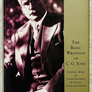 Puede incluir: Un libro de tapa dura titulado "The Basic Writings of C.G. Jung". La portada presenta un retrato de un hombre con traje y gafas. El lomo del libro es de color verde oliva claro con texto blanco. Editado con una introducción de Violet Staub de Laszlo.