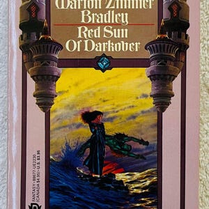 May include: A paperback book with the title "Red Sun of Darkover" by Marion Zimmer Bradley. The cover art depicts a fantasy scene with a woman and a creature by the sea. The author's name and title are visible.