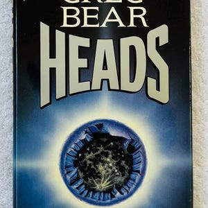 May include: A hardcover book titled "HEADS" by Greg Bear. The cover features the title in large, white, bold letters against a dark blue background. A blue sphere with a bright center is in the lower portion.