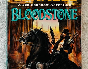 Op de afbeelding: Boekomslag van "Bloodstone" door David Gemmell. Het toont een man te paard, met een geweer, tegen een woestijnachtergrond. De titel en de naam van de auteur zijn prominent aanwezig.