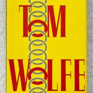 May include: A book cover with a vibrant yellow backdrop, featuring the name "TOM WOLFE" in bold, red, sans-serif lettering. The letters are intersected by a chain of interlocking black and white circles.
