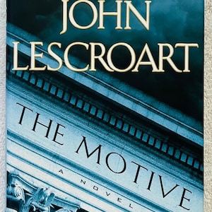 May include: A book cover for "The Motive" by John Lescroart. The title and author's name are in large, gold-colored letters against a dark blue background. The cover features a stylized image of a building facade.