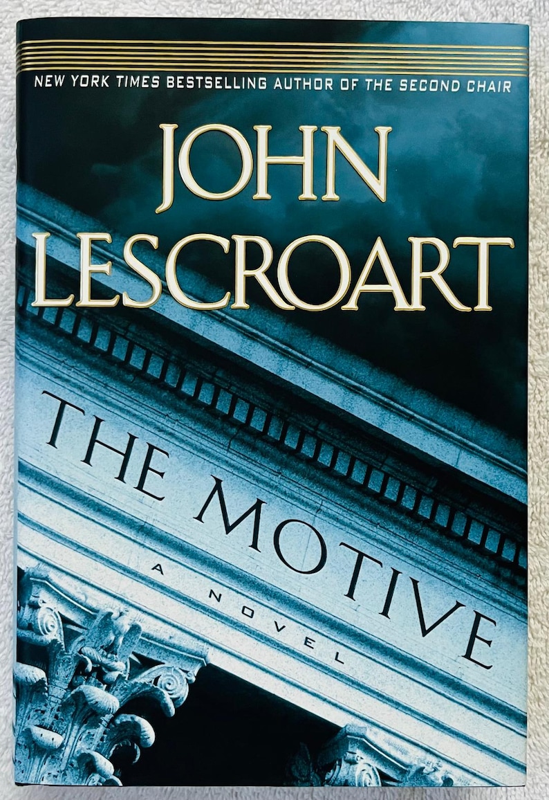 May include: A book cover for "The Motive" by John Lescroart. The title and author's name are in large, gold-colored letters against a dark blue background. The cover features a stylized image of a building facade.