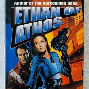 May include: A science fiction book cover featuring the title "ETHAN OF ATHOS" in blue and white lettering. The author's name, Lois McMaster Bujold, is at the top. The cover art depicts a woman in a blue outfit holding a futuristic weapon.