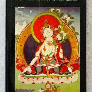 Pode incluir: Um livro de capa dura preto intitulado "Karma/Nirvana: Two Buddhist Tales by Paul Carus" com prefácio de Leo Tolstoy. A capa apresenta uma ilustração colorida de uma figura sentada com um halo, rodeada por elementos florais.