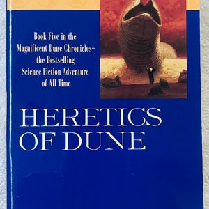 May include: A hardcover book titled "Heretics of Dune" by Frank Herbert. The cover is blue with white text and an illustration of a worm-like creature. The text reads "The New York Times Bestseller" and "Book Five in the Magnificent Dune Chronicles".