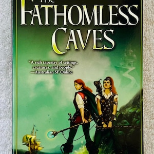 Op de afbeelding: Boekomslag voor "The Fathhomless Caves" van Kate Forsyth. De cover toont twee vrouwen in fantasy-kleding, een schip en een kasteel. Het boek maakt deel uit van de serie "The Witches of Eileanan".