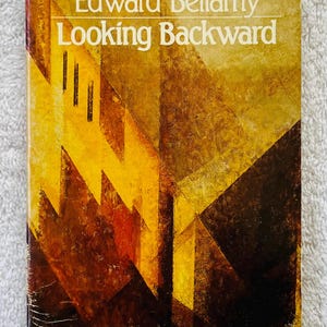 May include: A vintage paperback book titled "Looking Backward" by Edward Bellamy. The cover displays an abstract geometric design in yellow, brown, and black. The book is part of the Signet Classic series.