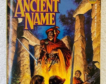 May include: A vintage paperback book titled "A Strange and Ancient Name" by Josepha Sherman. The cover art features a fantasy scene with figures holding swords, set against a backdrop of stone structures and a full moon. The book is a fantasy novel.